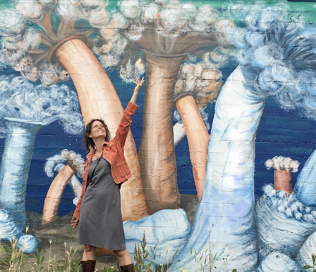 Carrie Ziegler stands in front of the blue mural she painted, smiling and triumphantly stretching her hand towards the sky.