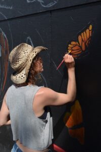 Carrie paints the wing of a monarch butterfly onto a mural outside. 