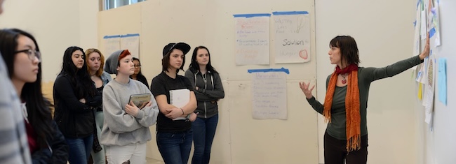 Carrie stands in a classroom, talking to a group of high school students. Brainstorming posters about hunger are posted on the walls around them.