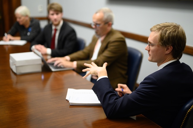 A white man sits at a conference room style table, with papers in front of him, gesturing as he speaks. Three other people in business attire sit in the seats next to him.