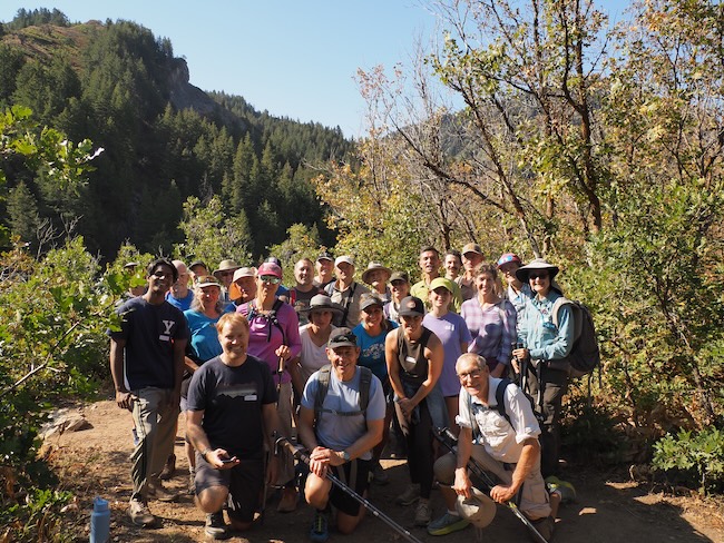 A group of 20+ people in hiking gear smiles at the camera, with forested mountains visible in the background