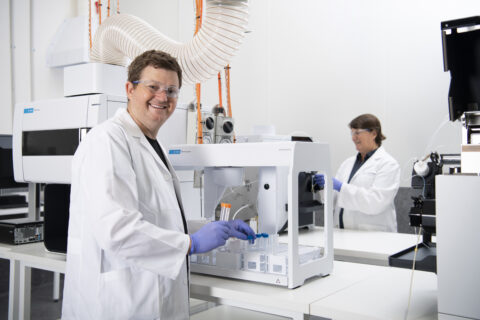 A white man in a white lab coat holds a test tube in a laboratory tracing energy transition minerals back to the mine